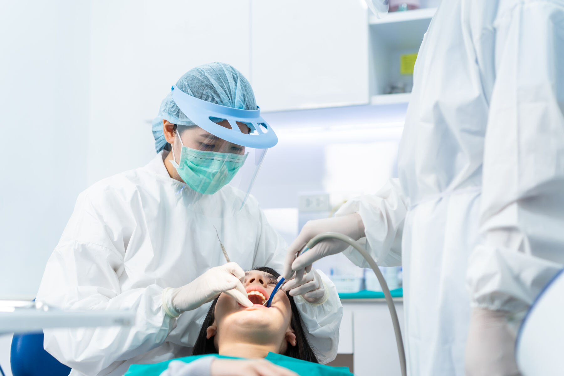 Asian expert dentist doctor wear PPE and mask examine patient's teeth at dental clinic. Dental assistant use suction to keep young girl's mouth dry due treatment procedures during Coronavirus pandemic